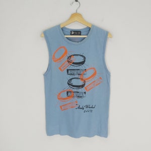 Puede incluir: Una camiseta sin mangas de color azul claro con un estampado gráfico de la obra Hamburger de Andy Warhol. La impresión presenta varias hamburguesas naranjas y negras con la palabra "Hamburger" impresa en naranja.