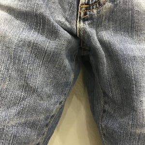 May include: Close-up of light blue denim jeans. The jeans show signs of wear and fading, with visible stitching in yellow thread. The image focuses on the front of the jeans, highlighting the texture and details of the fabric.