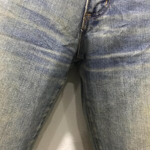 以下が含まれることがあります： ライトブルーのデニムジーンズのクローズアップ。ジーンズは、生地全体に明るい水平ストライプのある色あせたウォッシュ効果を示しています。画像は、ウエストバンドと脚の一部を含む上部に焦点を当てています。