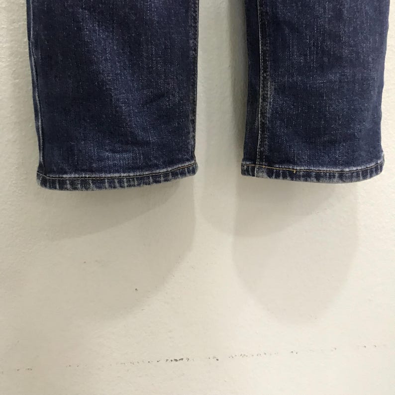May include: Close-up of the hem of a pair of dark blue denim jeans. The jeans have a classic, straight-leg cut with a slightly worn appearance. The hems are neatly stitched.