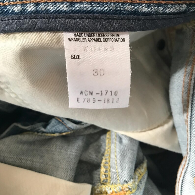 K&ouml;nnte beinhalten: Nahaufnahme eines wei&szlig;en Etiketts in einer blauen Jeans. Das Etikett lautet "MADE UNDER LICENSE FROM WRANGLER APPAREL CORPORATION" und "GR&Ouml;SSE 30". Die Jeans ist hellblau mit sichtbaren N&auml;hten.