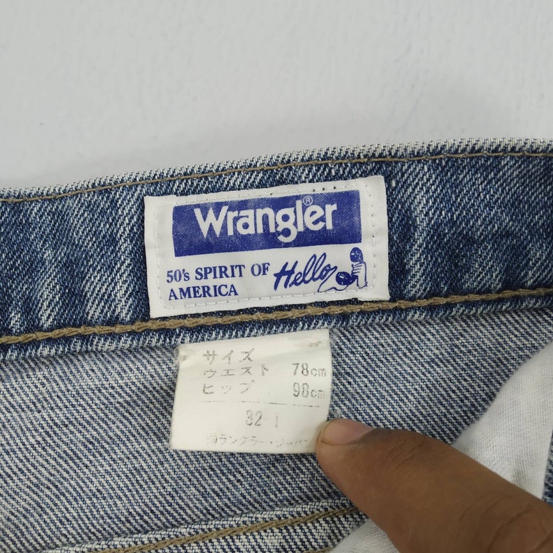 Może przedstawiać: Zbliżenie niebieskich dżins&oacute;w z metką Wrangler. Na metce widnieje napis "Wrangler 50's Spirit of America Hello". Dżinsy mają metkę z rozmiarem z wymiarami w centymetrach. Talia ma 78 cm, a biodra 98 cm.
