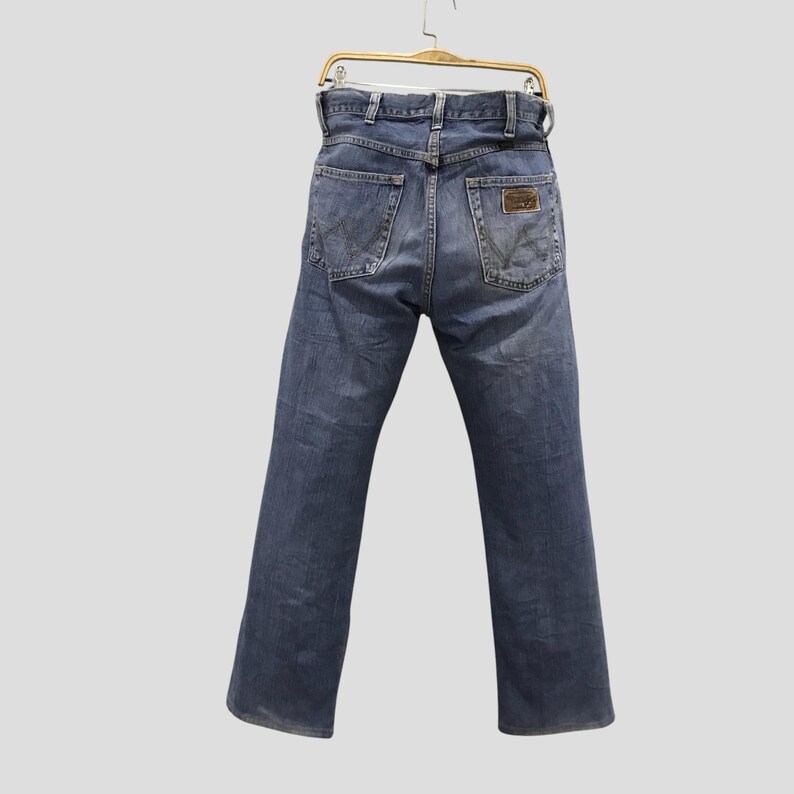 May include: Faded blue denim jeans with a classic five-pocket design. The jeans feature a straight leg cut and a brown leather patch on the back pocket. The jeans are hanging on a gold hanger.