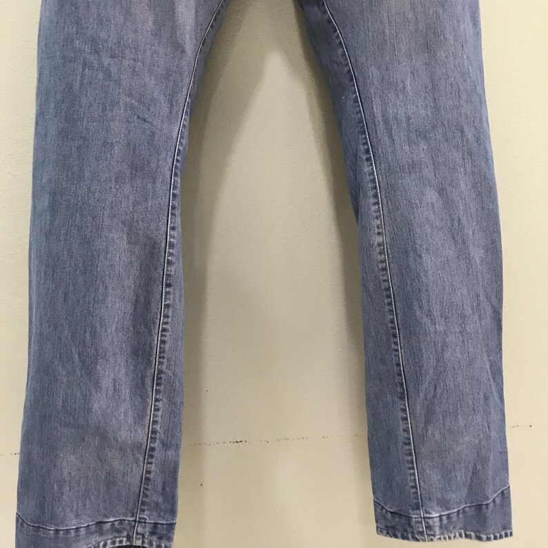 May include: A pair of blue denim jeans featuring a straight-leg cut with visible vertical seams running down the front of each leg. The denim has a medium-wash finish with subtle fading throughout the fabric and a horizontal hem at the bottom.