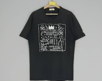 May include: Black t-shirt featuring a white graphic print of a drawing by artist Jean-Michel Basquiat. The drawing includes the text "Test Pressing, Version One, Volume One,  Downtown Record Co., New York, N.Y., U.S.A."