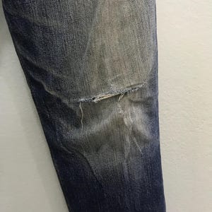 May include: Close-up of a section of blue denim jeans with a distressed knee. The denim has faded areas and a visible tear at the knee, revealing the fabric's frayed edges. The jeans are against a white background.