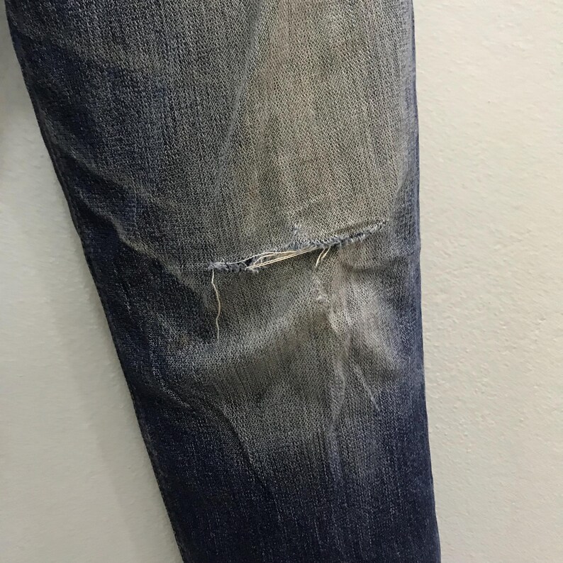 May include: Close-up of a section of blue denim jeans with a distressed knee. The denim has faded areas and a visible tear at the knee, revealing the fabric's frayed edges. The jeans are against a white background.