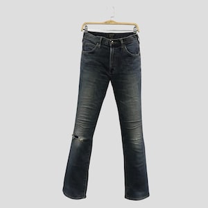 Pu&ograve; includere: Jeans in denim blu scuro con un dettaglio strappato sul ginocchio, appesi a una gruccia di legno. I jeans hanno un classico design a cinque tasche e una chiusura con bottone e cerniera. I jeans sono leggermente sbiaditi.