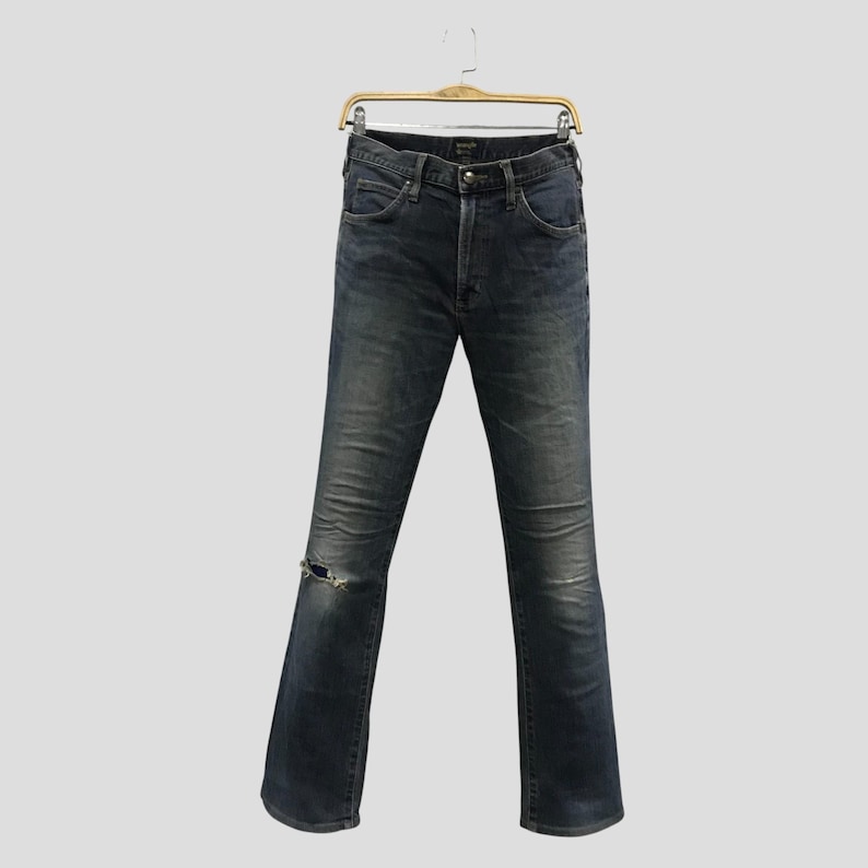 Pu&ograve; includere: Jeans in denim blu scuro con un dettaglio strappato sul ginocchio, appesi a una gruccia di legno. I jeans hanno un classico design a cinque tasche e una chiusura con bottone e cerniera. I jeans sono leggermente sbiaditi.