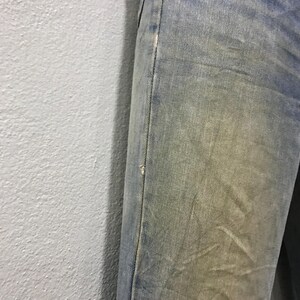 以下が含まれることがあります： 色あせた青色のデニムジーンズのクローズアップ。ジーンズは、わずかなダメージと微妙な黄ばみ効果で、使用感を示しています。縫い目はフレームの右側にあり、膝の近くに小さな破れがあります。生地は質感のある外観をしています。