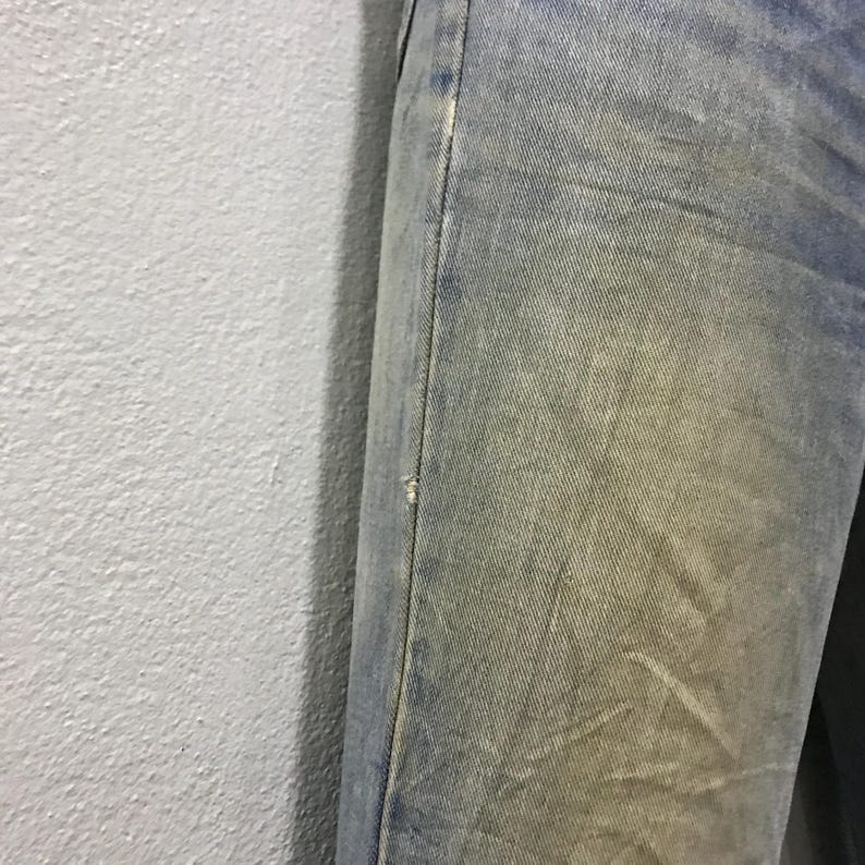 以下が含まれることがあります： 色あせた青色のデニムジーンズのクローズアップ。ジーンズは、わずかなダメージと微妙な黄ばみ効果で、使用感を示しています。縫い目はフレームの右側にあり、膝の近くに小さな破れがあります。生地は質感のある外観をしています。