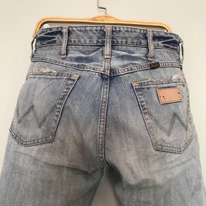 K&ouml;nnte beinhalten: Hellblaue Jeans aus Denim mit einem Used-Look, mit dem Wrangler-Logo auf der Ges&auml;&szlig;tasche. Die Jeans h&auml;ngen an einem Holzb&uuml;gel und zeigen die Ges&auml;&szlig;taschen und die klassische 'W'-Naht.
