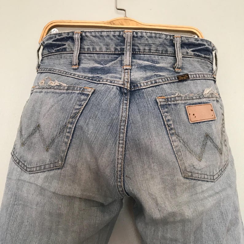 K&ouml;nnte beinhalten: Hellblaue Jeans aus Denim mit einem Used-Look, mit dem Wrangler-Logo auf der Ges&auml;&szlig;tasche. Die Jeans h&auml;ngen an einem Holzb&uuml;gel und zeigen die Ges&auml;&szlig;taschen und die klassische 'W'-Naht.