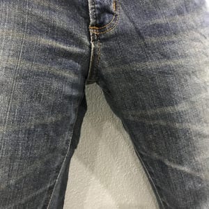 Pu&ograve; includere: Primo piano di jeans di denim blu. I jeans hanno un classico design a cinque tasche con chiusura con bottone e cerniera. Il denim ha un aspetto sbiadito e usurato con sottili variazioni di colore.