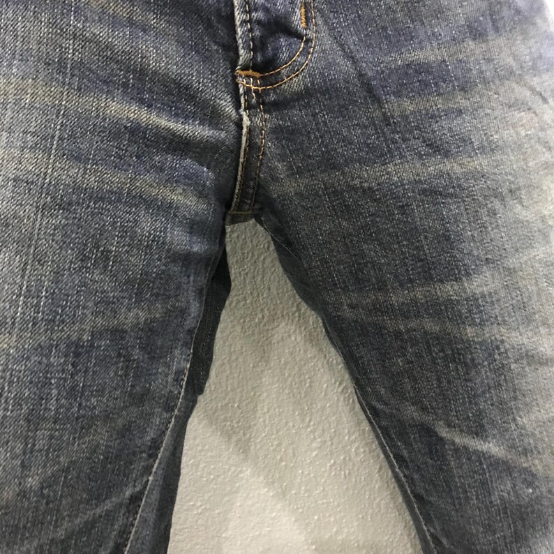 Pu&ograve; includere: Primo piano di jeans di denim blu. I jeans hanno un classico design a cinque tasche con chiusura con bottone e cerniera. Il denim ha un aspetto sbiadito e usurato con sottili variazioni di colore.