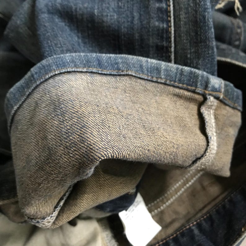 Pu&ograve; includere: Primo piano di un paio di jeans blu, con il risvolto arrotolato. L'interno del risvolto rivela un tessuto denim pi&ugrave; chiaro e sbiadito. Le cuciture sono visibili lungo le cuciture e un'etichetta bianca &egrave; parzialmente visibile.