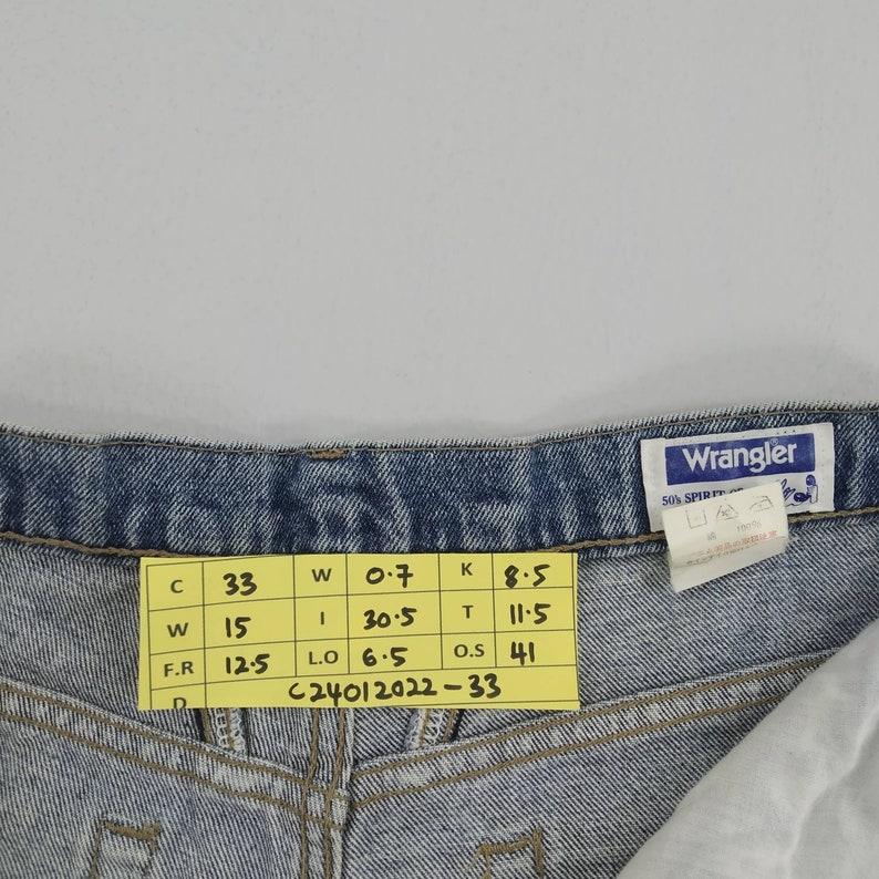 Może przedstawiać: Zbliżenie jasnoniebieskich dżins&oacute;w Wrangler. Ż&oacute;łta etykieta wyświetla wymiary: C 33, W 15, F.R 12,5 cala (31,75 cm). Dżinsy mają klasyczny pięciokieszeniowy kr&oacute;j i metkę Wrangler.