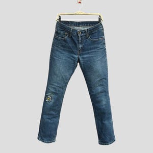 Könnte beinhalten: Blaue Jeans aus Denim mit verwaschener Optik, hängen an einem Kleiderbügel. Die Jeans haben einen dekorativen Blumenfleck am linken Knie. Die Jeans haben ein klassisches Fünf-Taschen-Design und einen Knopf- und Reißverschluss.