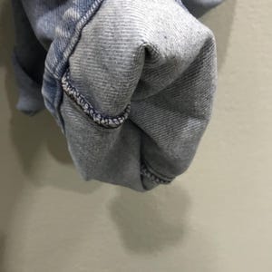 May include: Close-up of light blue denim fabric, showing the folded hem of a pair of jeans. The hem is turned up, revealing the inside of the denim. The stitching is visible along the edge of the fold.