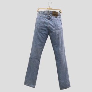 May include: Light blue denim jeans hanging on a gold hanger. The jeans have a classic five-pocket design, a brown leather patch, and a straight leg. The back pockets have a decorative stitch.