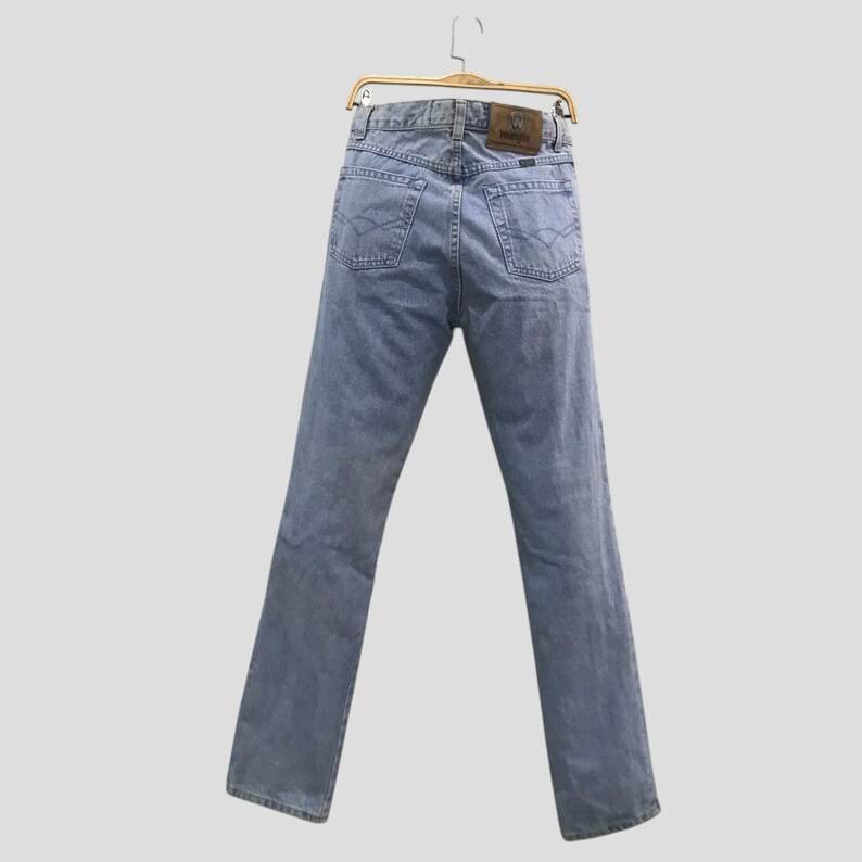 May include: Light blue denim jeans hanging on a gold hanger. The jeans have a classic five-pocket design, a brown leather patch, and a straight leg. The back pockets have a decorative stitch.