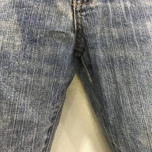 Op de afbeelding: Close-up van een blauwe spijkerbroek. De jeans heeft een klassiek ontwerp met vijf zakken, met zichtbare stiksels en een knoop- en ritssluiting. De denim heeft een vervaagde, versleten uitstraling.
