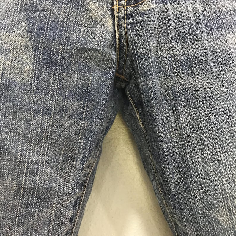 Op de afbeelding: Close-up van een blauwe spijkerbroek. De jeans heeft een klassiek ontwerp met vijf zakken, met zichtbare stiksels en een knoop- en ritssluiting. De denim heeft een vervaagde, versleten uitstraling.