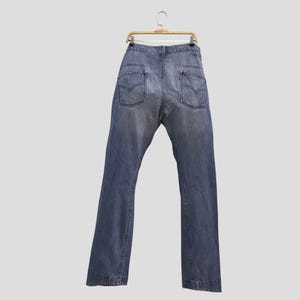 May include: A pair of faded blue denim jeans hanging on a wire hanger. The jeans feature a classic five-pocket design with two back pockets, each displaying a stitched V-shaped detail and a small red tab on the right pocket edge.