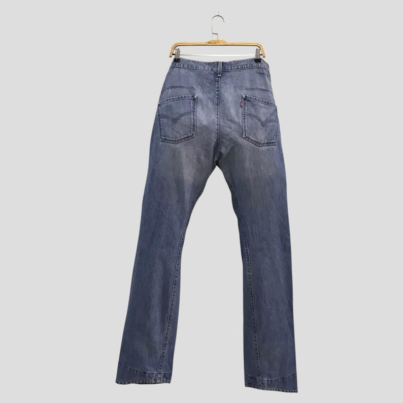 May include: A pair of faded blue denim jeans hanging on a wire hanger. The jeans feature a classic five-pocket design with two back pockets, each displaying a stitched V-shaped detail and a small red tab on the right pocket edge.