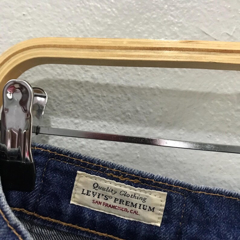 May include: Close-up of a pair of blue denim jeans with a white label that reads "Quality Clothing LEVI'S PREMIUM SAN FRANCISCO, CAL." The jeans are hanging on a wooden hanger.