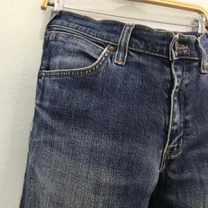 May include: Close-up of blue denim jeans. The jeans have a classic five-pocket design, with a button and zip closure. The denim has a faded, worn look, with visible stitching and rivets.