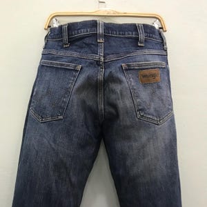 May include: Rear view of blue denim Wrangler jeans. The jeans feature two back pockets and a brown leather patch with the Wrangler logo. The jeans are suspended from a wooden clothes hanger.