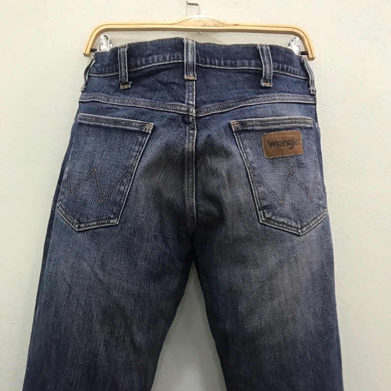 May include: Rear view of blue denim Wrangler jeans. The jeans feature two back pockets and a brown leather patch with the Wrangler logo. The jeans are suspended from a wooden clothes hanger.