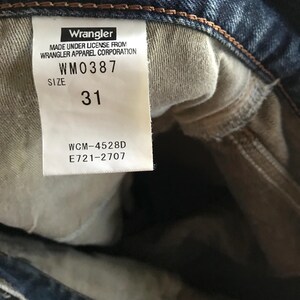 Pu&ograve; includere: Primo piano di un'etichetta di jeans Wrangler. L'etichetta bianca presenta testo nero, tra cui il marchio, il numero di modello WM0387, la taglia 31 e altri dettagli. I jeans sono in denim di lavaggio medio.