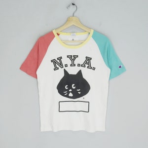 May include: White t-shirt with a black cat graphic and the text "N.Y.A." in black. The shirt has a red and blue colour block design on the sleeves.