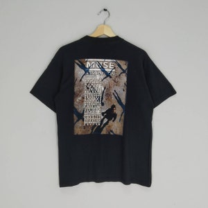 Puede incluir: Camiseta negra con un estampado gráfico de la banda Muse. El gráfico presenta el nombre de la banda, el título del álbum "Absolution" y una lista de fechas de la gira.