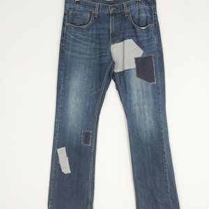 May include: Blue denim jeans with patches of gray and navy fabric. The jeans are hanging on a metal hanger against a white background. The jeans have a classic five-pocket design and a straight leg cut.