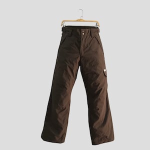 Op de afbeelding: Donkerbruine skibroek met een hoge taille, een knoopsluiting aan de voorzijde en een kleine witte logopatch op de zak op de linkerdij. Deze duurzame broek is gemaakt van een gladde, waterafstotende stof en heeft wijde broekspijpen.