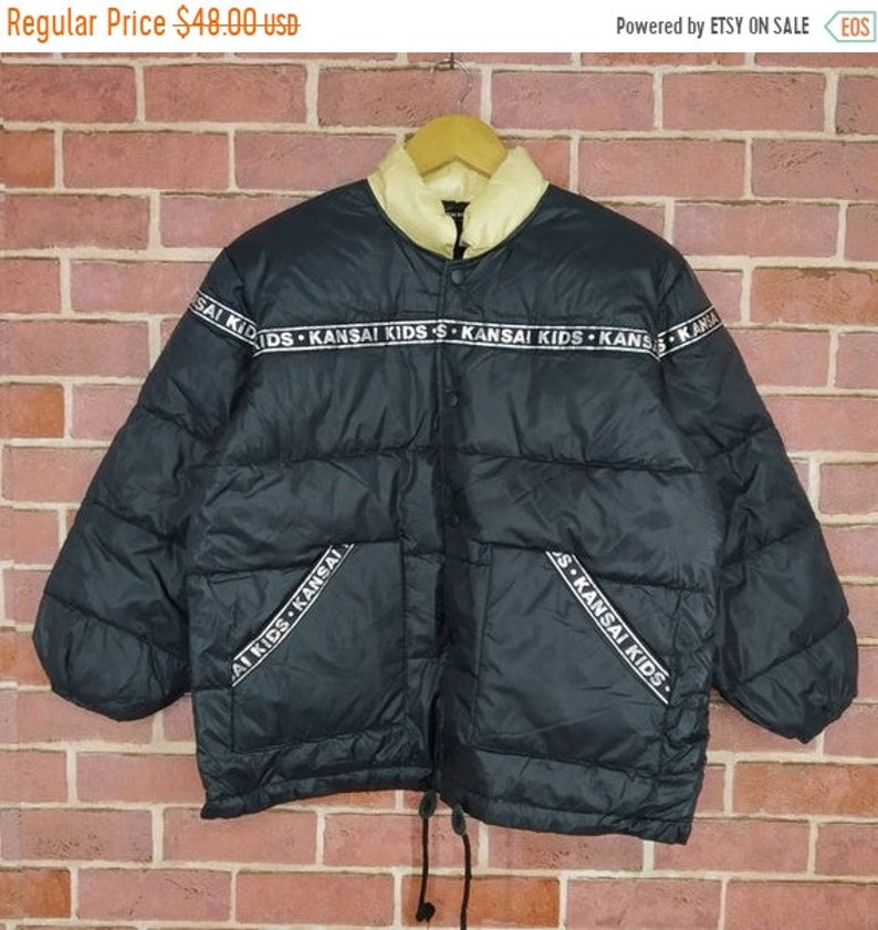 May include: Black puffer jacket with a white fleece lining and a zipper closure. The jacket has a band around the waist and the sleeves with the text "KANSAI KIDS" repeated.
