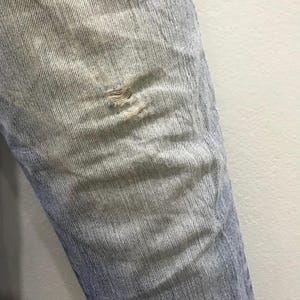 May include: Close-up of light blue denim jeans with a distressed, ripped knee. The denim shows fading and wear, with visible threads at the tear. The jeans have a classic five-pocket design and a straight leg.