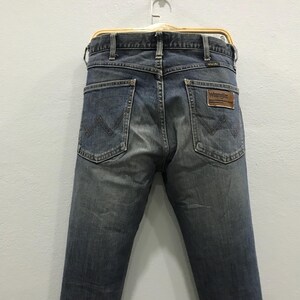Pu&ograve; includere: Jeans di denim blu sbiadito appesi a una gruccia di legno. I jeans hanno un classico design a cinque tasche, con il logo Wrangler su una toppa in pelle marrone. Le tasche posteriori presentano le cuciture "W" distintive di Wrangler.