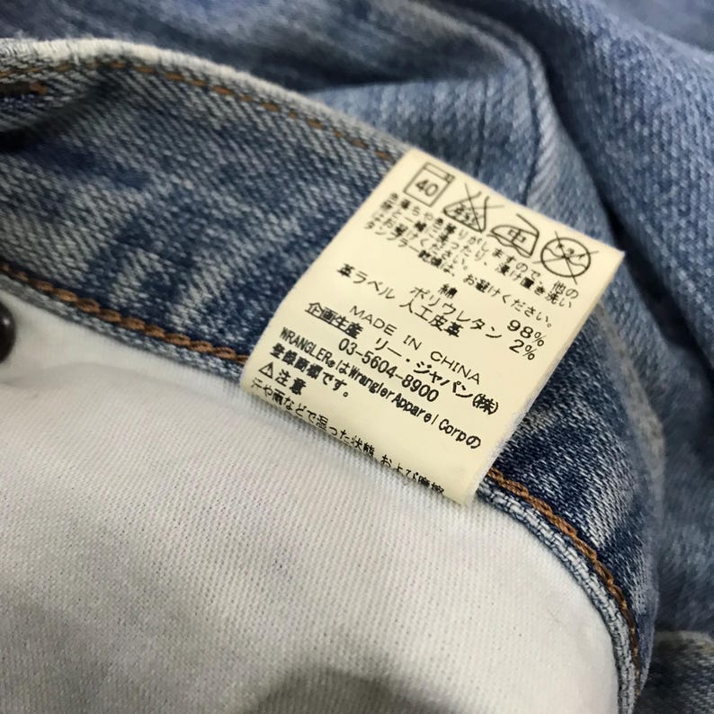 May include: Close-up of a light blue denim garment with a white interior. A rectangular tag is attached, displaying Japanese text and care symbols. The tag indicates the garment is made in China and lists the fabric composition.