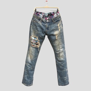 Może przedstawiać: Niebieskie dżinsy z przetarciami, w tym podarte łaty na lewym kolanie. Pas ma kwiatowy nadruk. Dżinsy wiszą na wieszaku na neutralnym tle.