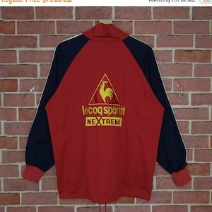 Puede incluir: Chaqueta de ch&aacute;ndal roja y negra con una raya blanca en cada manga. La chaqueta presenta un gran logotipo bordado de un gallo en un tri&aacute;ngulo con el texto "le coq sportif" y "NEXTREME" debajo.