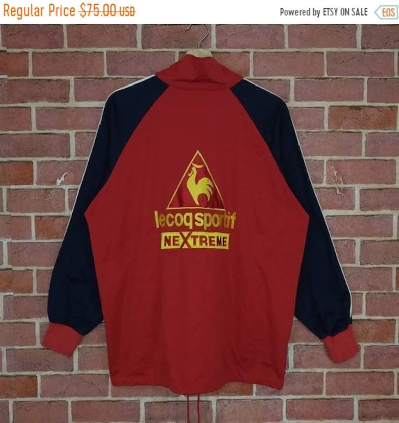 Puede incluir: Chaqueta de ch&aacute;ndal roja y negra con una raya blanca en cada manga. La chaqueta presenta un gran logotipo bordado de un gallo en un tri&aacute;ngulo con el texto "le coq sportif" y "NEXTREME" debajo.