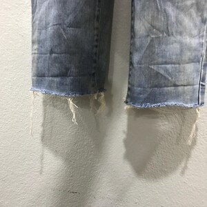 以下が含まれることがあります： ライトブルーデニムジーンズのほつれた裾のクローズアップ。ジーンズは、目に見える摩耗や損傷があり、ダメージ加工が施されており、端は意図的に未完成で、糸がほつれています。