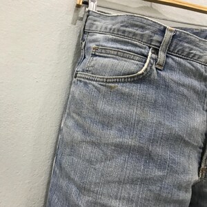 May include: Light blue denim jeans, featuring a classic five-pocket design. The jeans are faded, showing signs of wear, and are hanging on a wooden hanger. The close-up shot highlights the texture and details of the fabric.