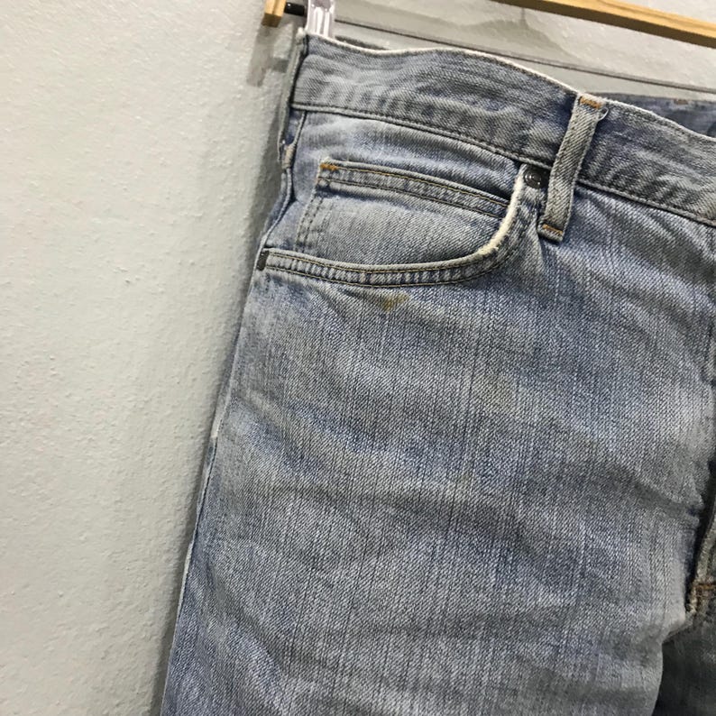 May include: Light blue denim jeans, featuring a classic five-pocket design. The jeans are faded, showing signs of wear, and are hanging on a wooden hanger. The close-up shot highlights the texture and details of the fabric.