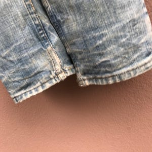 May include: Close-up of light blue denim shorts with distressed detailing. The shorts show signs of wear and tear, with frayed edges and faded patches. The fabric has a textured appearance.