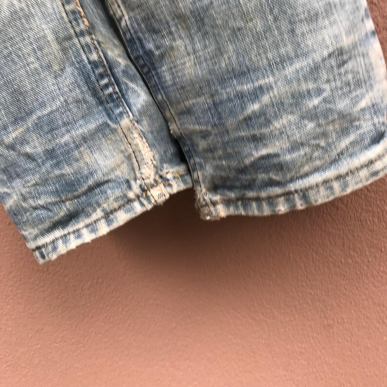 May include: Close-up of light blue denim shorts with distressed detailing. The shorts show signs of wear and tear, with frayed edges and faded patches. The fabric has a textured appearance.